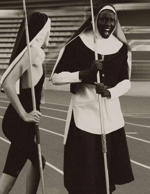 Two adult women in religious nun costumes on a running track, one with an open mouth, holding javelins, indoor setting.