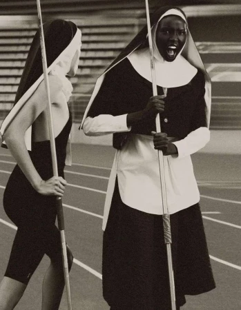 Two adult women in religious nun costumes on a running track, one with an open mouth, holding javelins, indoor setting.