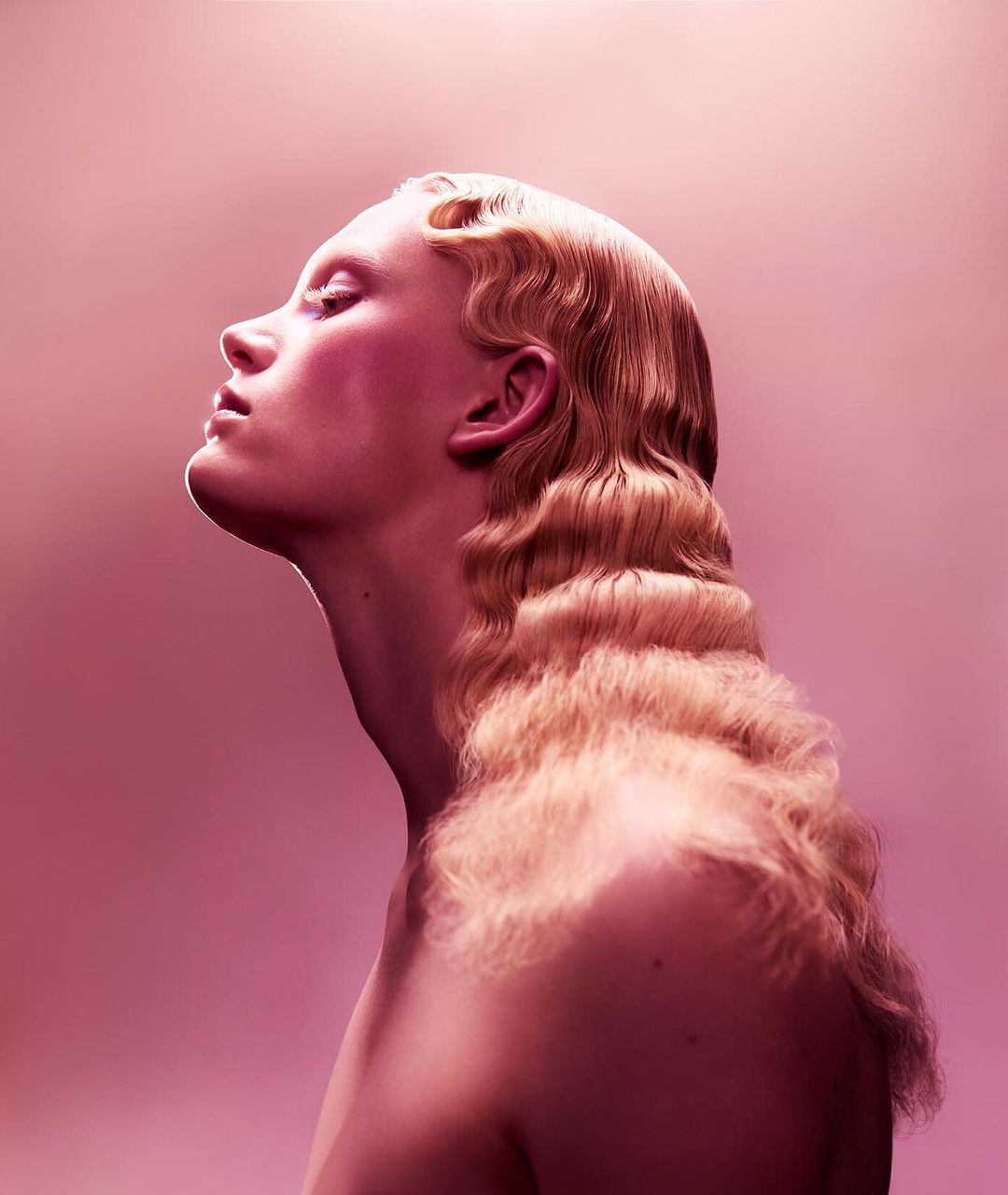 Profile portrait of a pale-skinned adult female with blonde wavy hair, looking upward against a graduated pink background in studio lighting.