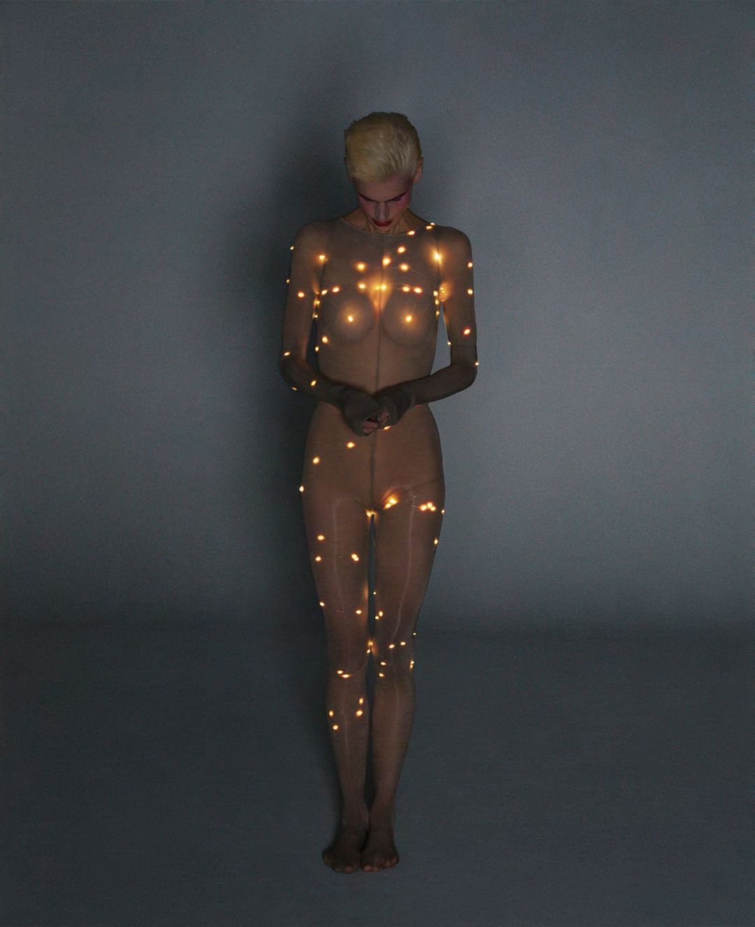Full-length portrait of a slender, light-skinned adult woman covered in tiny string lights, set against a neutral gray background in studio lighting.