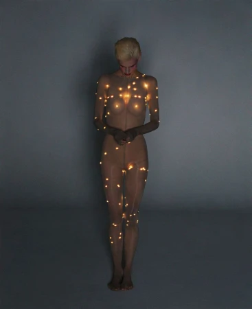 Full-length portrait of a slender, light-skinned adult woman covered in tiny string lights, set against a neutral gray background in studio lighting.