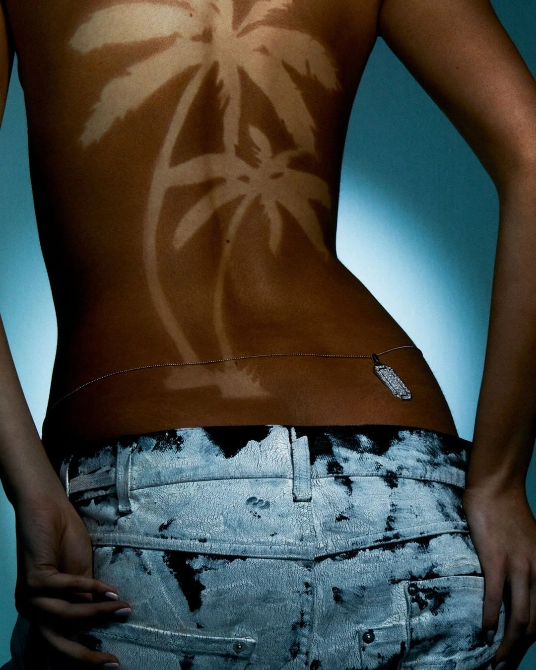 Woman with a palm tree sun tan line on her back wearing patterned light-wash jeans against a blue gradient background.