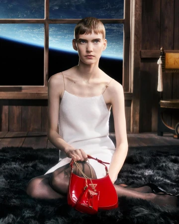 Model with short hair wearing a white slip dress seated on a black fur rug holding a red patent handbag