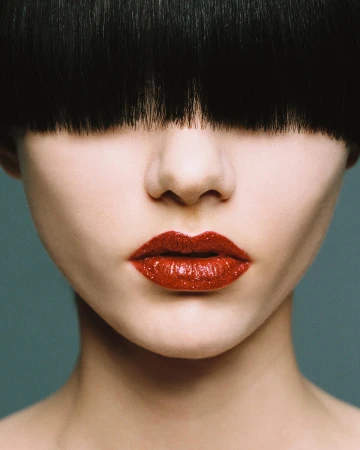 Extreme close-up portrait of a woman with heavy black blunt bangs covering her eyes and glossy red glitter lipstick.