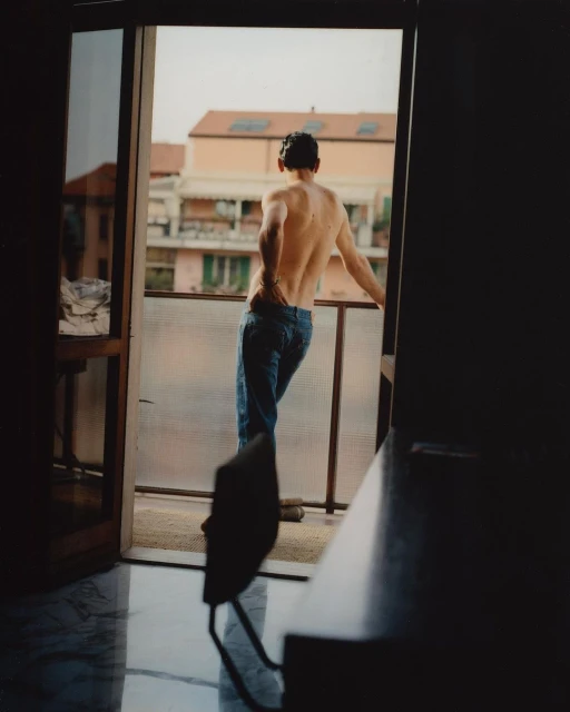 Back view of a shirtless man in jeans leaning on a balcony railing, overlooking a European cityscape on a sunny day.