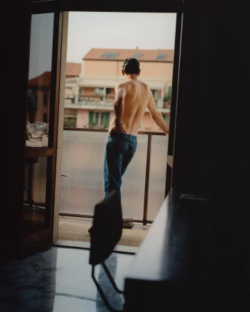 Back view of a shirtless man in jeans leaning on a balcony railing, overlooking a European cityscape on a sunny day.