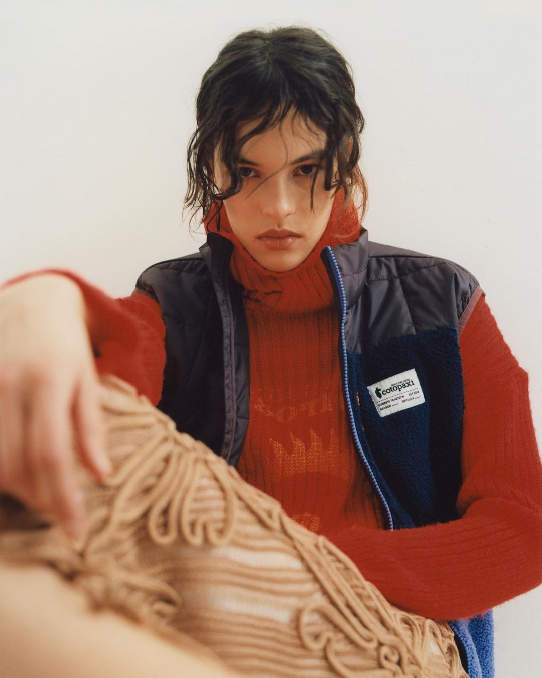 A young adult female poses indoors wearing a red turtleneck, black vest, and beige crocheted fabric. The image is shot in a photo studio.