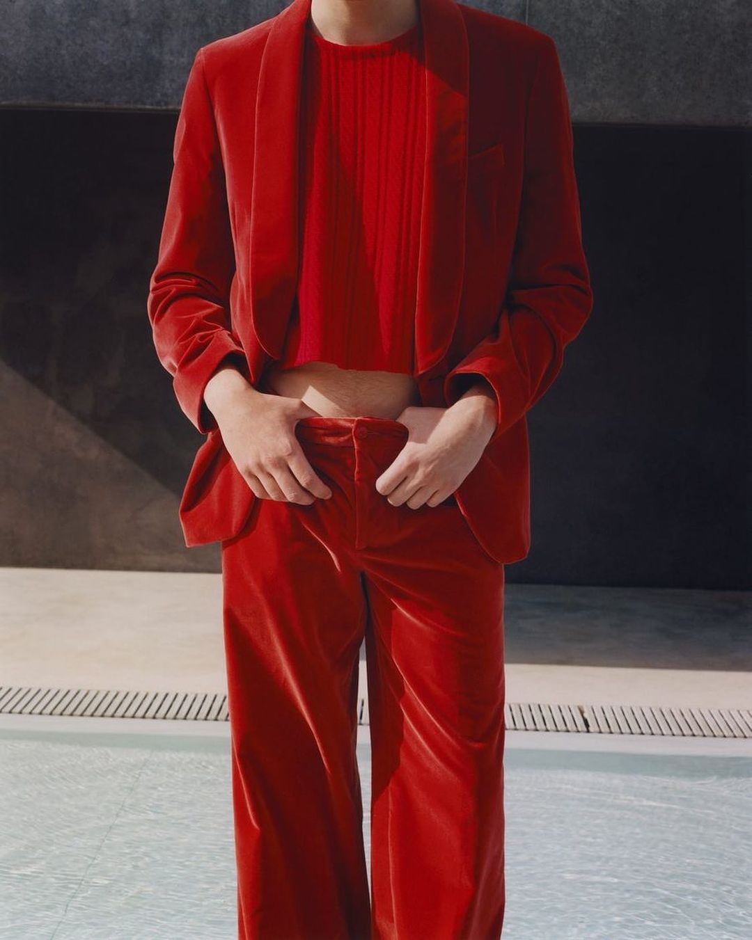Person in a red velvet suit and top stands near a pool, creating a bold fashion statement with a blend of formal and casual styles.