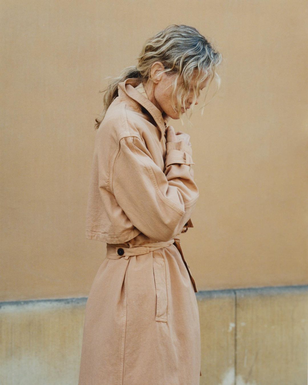 Woman in beige trench coat stands against a neutral wall. The model's expression is downcast, her hands clasped. Outdoor natural lighting.