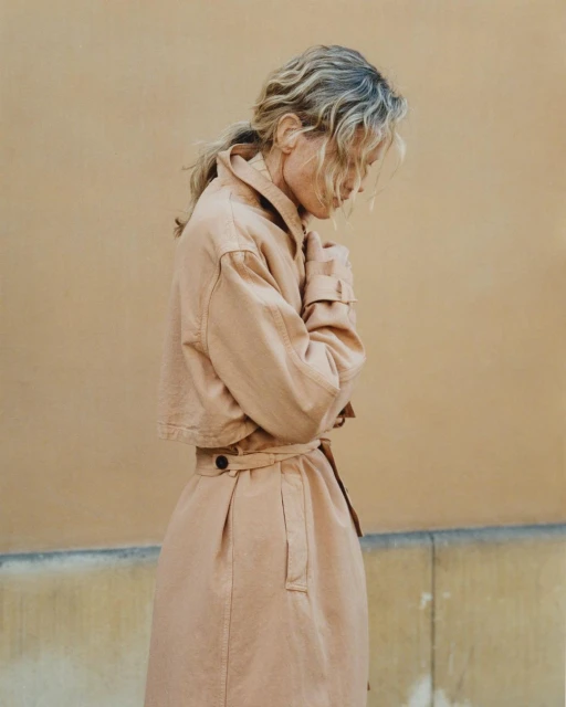 Woman in beige trench coat stands against a neutral wall. The model's expression is downcast, her hands clasped. Outdoor natural lighting.
