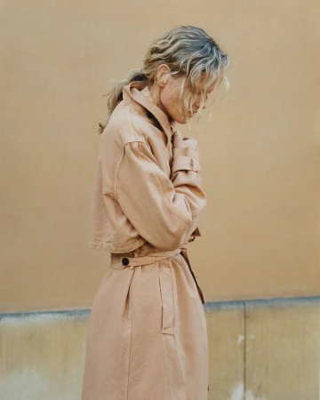 Woman in beige trench coat stands against a neutral wall. The model's expression is downcast, her hands clasped. Outdoor natural lighting.
