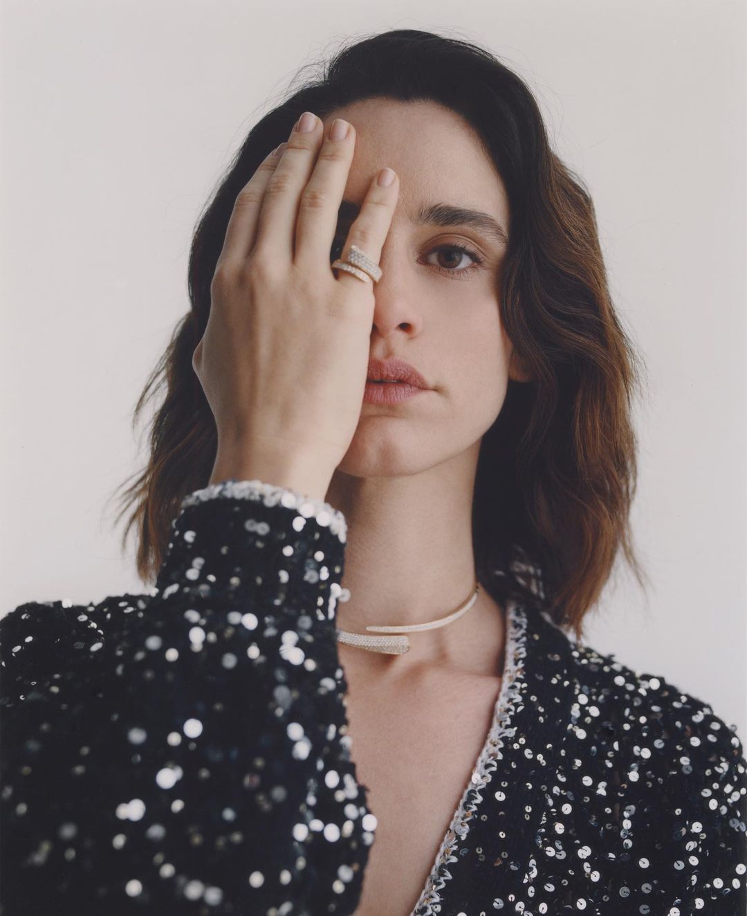 Close-up portrait of a brunette woman with hand over her eye, wearing a black sequined jacket and modern jewellery against a white backdrop.