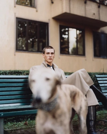 A young man seated on a green park bench wearing a long coat, with a blurred dog in the foreground.
