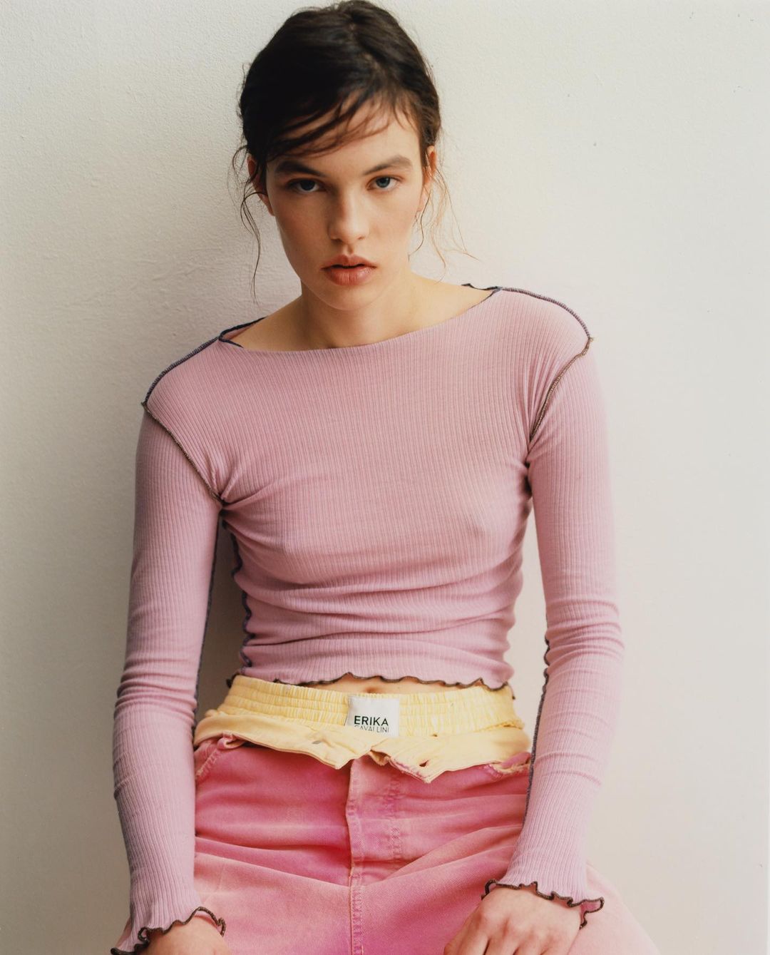 A young woman poses indoors wearing a pink ribbed long-sleeve top and pink jeans, looking at the camera with a neutral facial expression.