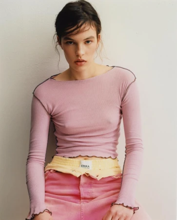 A young woman poses indoors wearing a pink ribbed long-sleeve top and pink jeans, looking at the camera with a neutral facial expression.