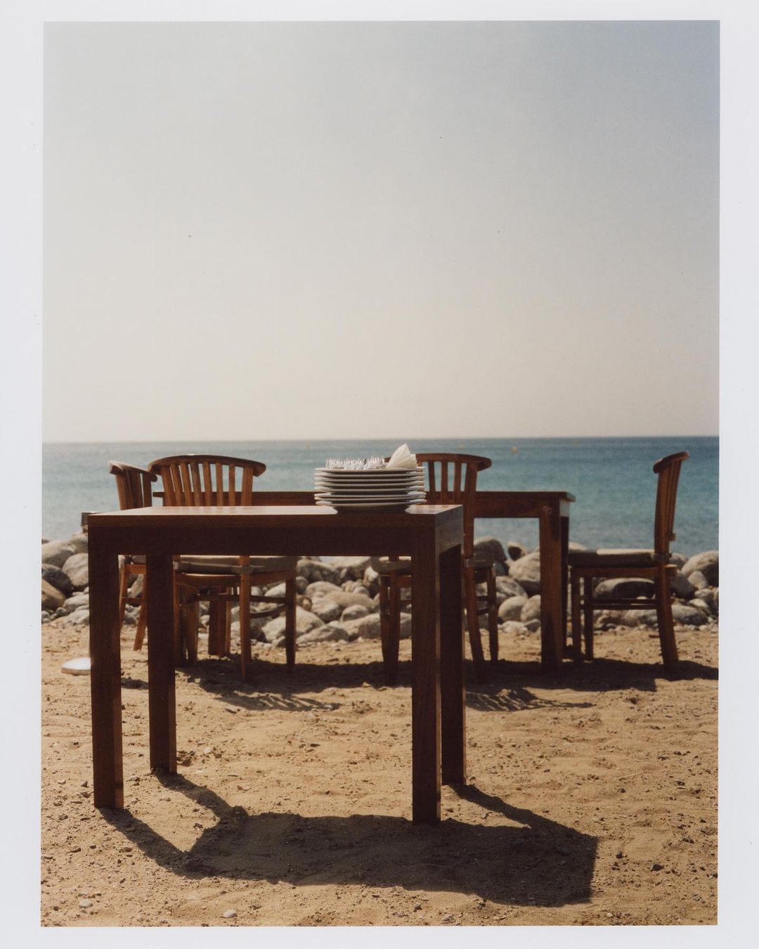 Ocean view with wooden tables and chairs on a sandy beach. Stacked white plates suggest a restaurant or cafe setting near the water.