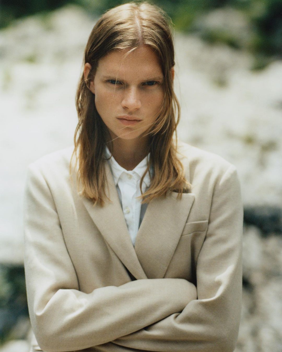 Portrait of a young adult woman with long blonde hair, wearing a beige suit and white shirt, standing outdoors against a rock backdrop.