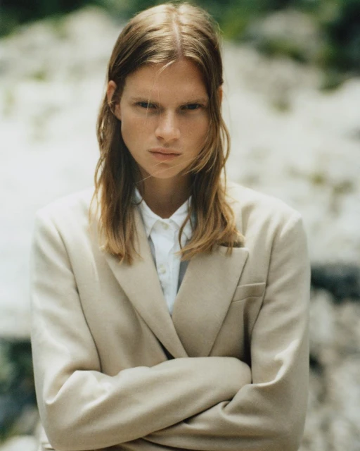 Portrait of a young adult woman with long blonde hair, wearing a beige suit and white shirt, standing outdoors against a rock backdrop.