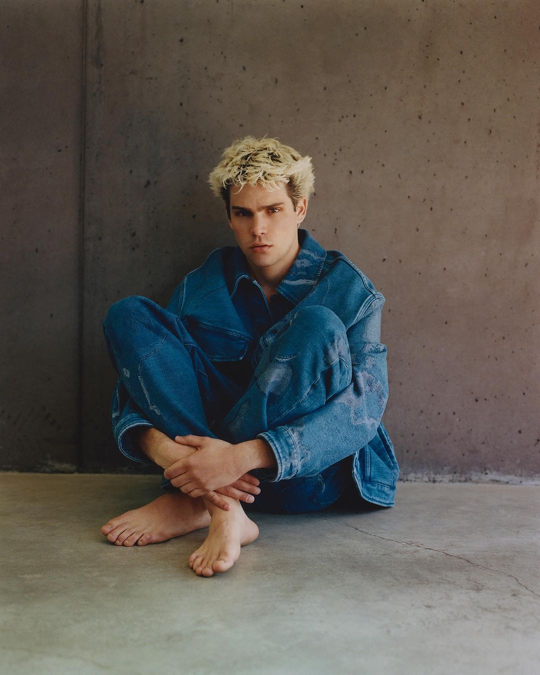Young man with blonde hair wearing a blue denim jacket and matching jeans seated barefoot against a concrete wall