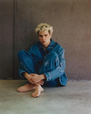 Young man with blonde hair wearing a blue denim jacket and matching jeans seated barefoot against a concrete wall