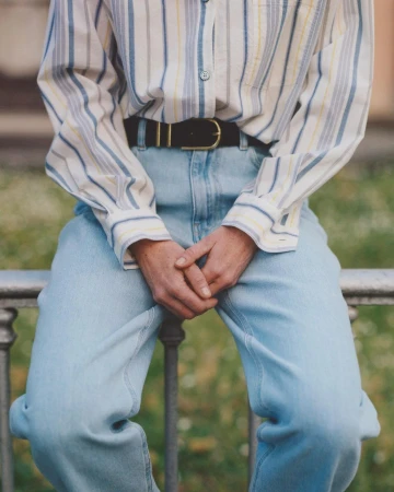 Person seated wearing a striped button-down shirt, black belt, and light blue jeans, photographed by Carlo Piro