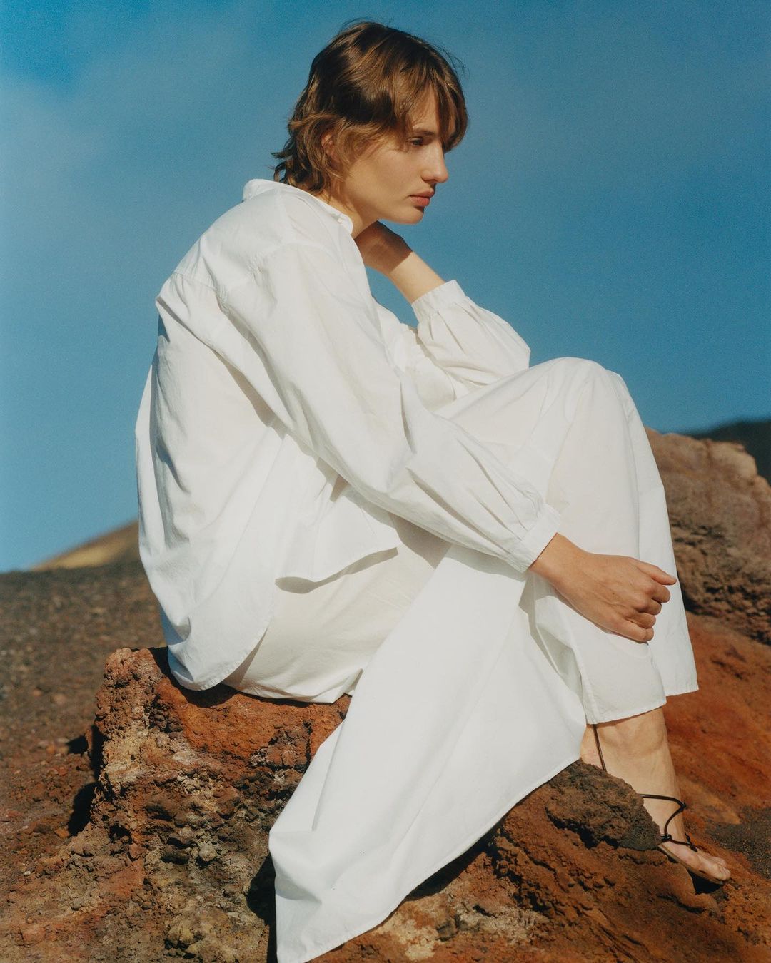 Fashion shot of woman in white outfit seated on rock formation against blue sky, promoting clothing design with natural backdrop.