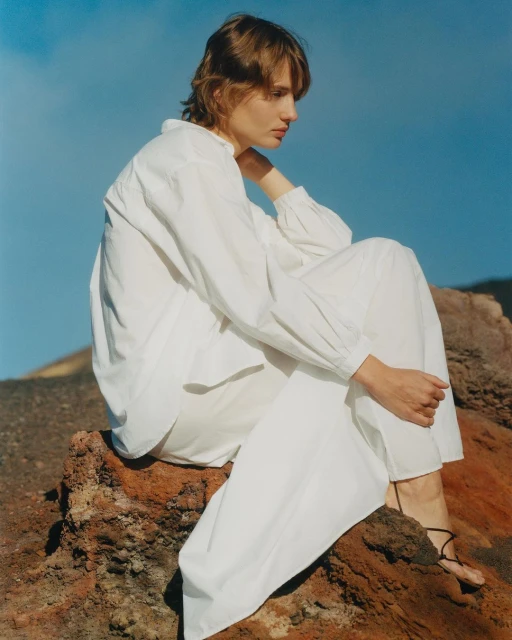 Fashion shot of woman in white outfit seated on rock formation against blue sky, promoting clothing design with natural backdrop.