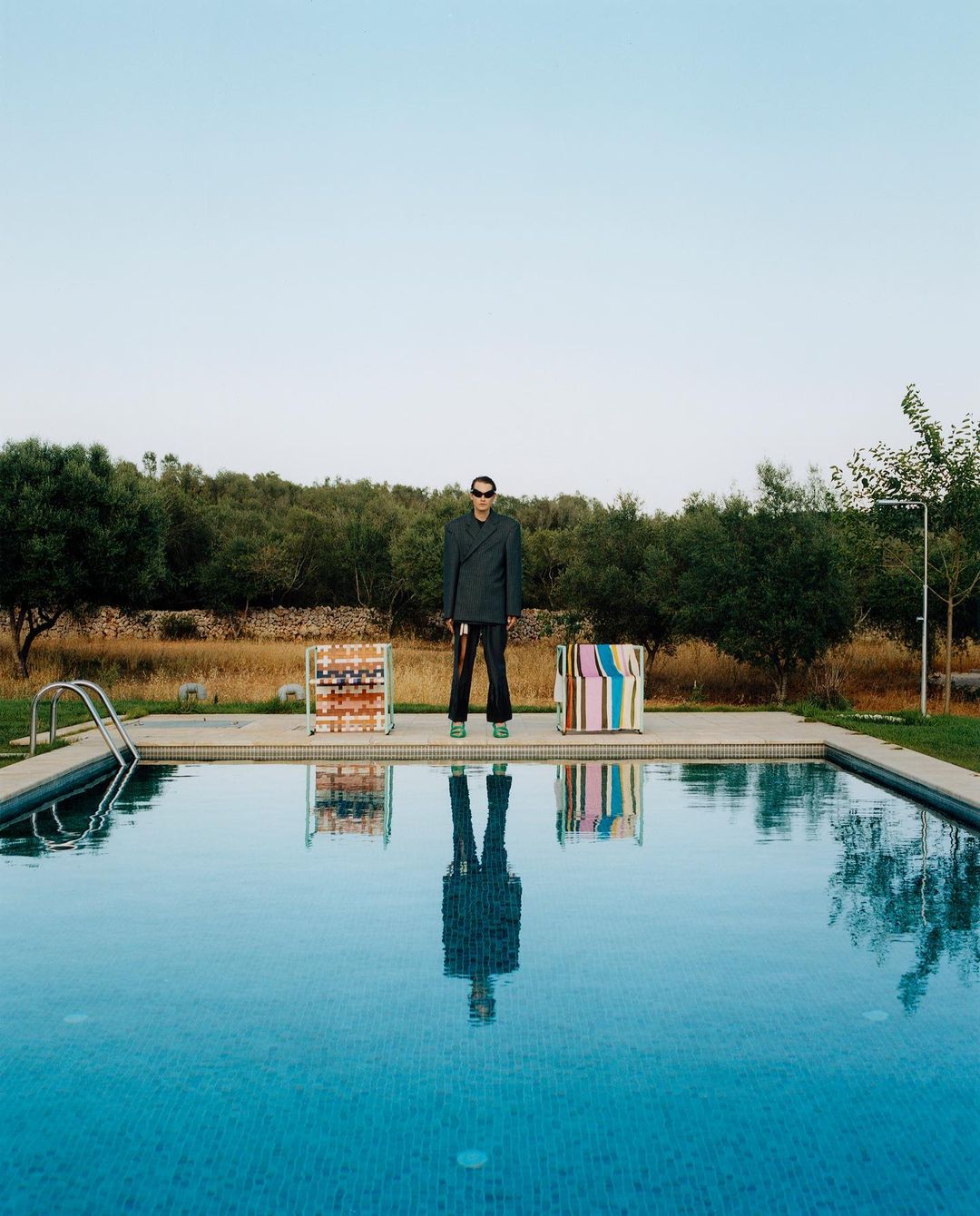 A man in a suit stands by a swimming pool with luggage, reflecting in the water, set against a backdrop of lush greenery and a clear sky.