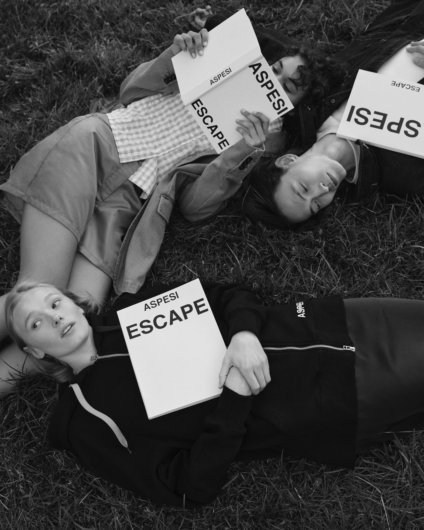 Monochrome image of four models lying on grass holding ASPESI ESCAPE branded books, creating a visually cohesive brand message.