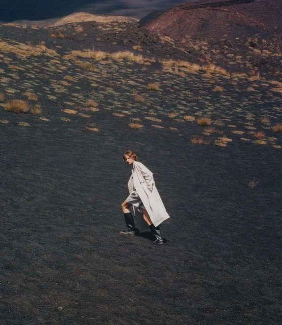 Woman in a cream coat and black boots walks across a black volcanic landscape with sparse vegetation under an overcast sky.