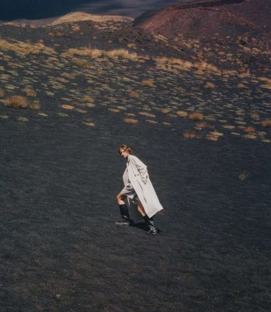 Woman in a cream coat and black boots walks across a black volcanic landscape with sparse vegetation under an overcast sky.