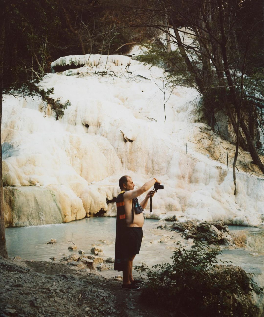 Adult male stands near natural waterfall, holding camera. Outdoor shot with trees, rocks, and water cascading down rocks.