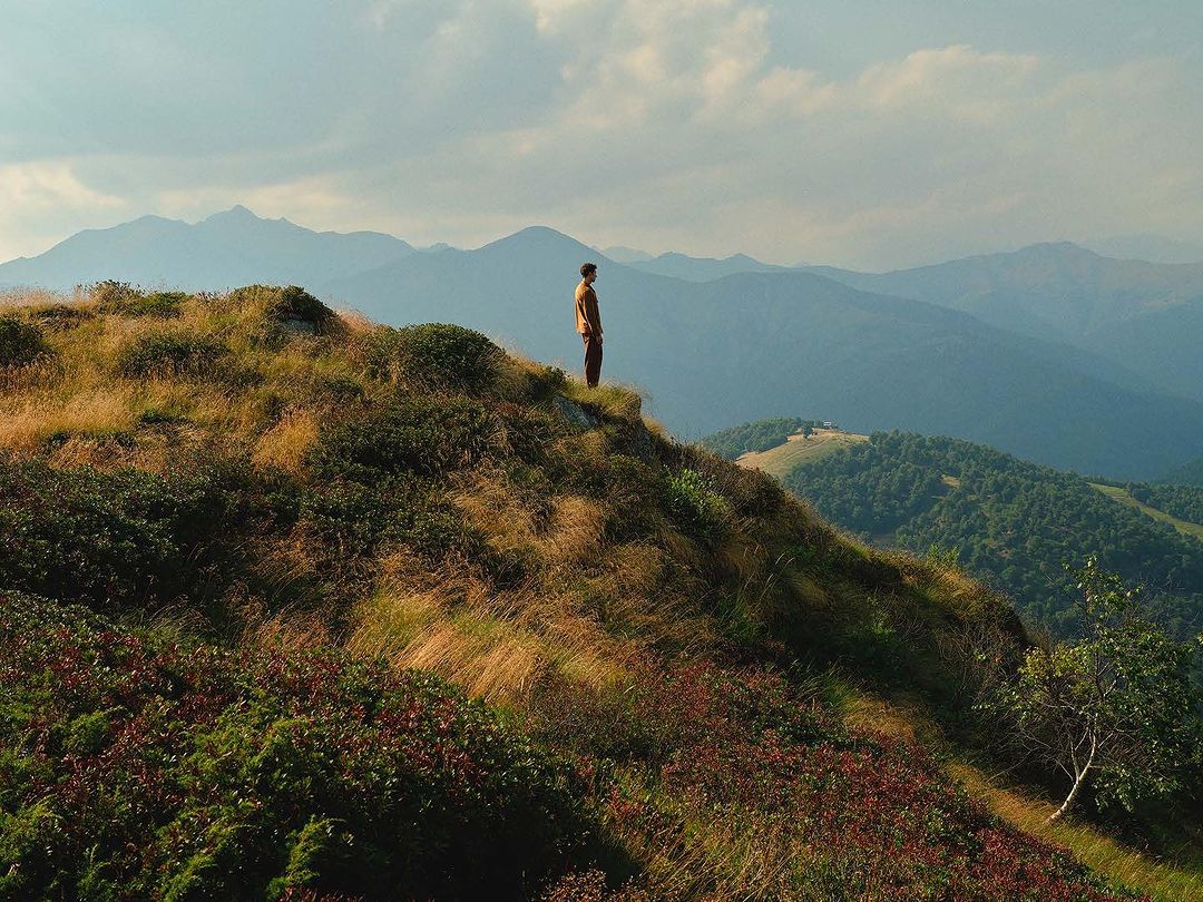 A lone adult stands on a grassy hilltop with a view of distant mountains under a soft, cloudy sky, creating a serene landscape.