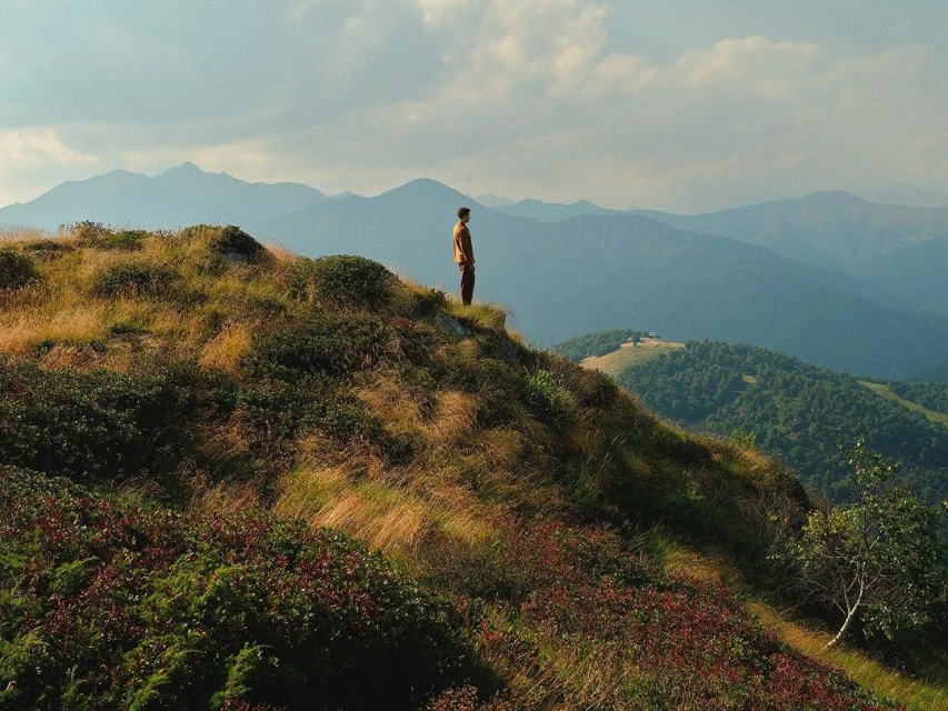 A lone adult stands on a grassy hilltop with a view of distant mountains under a soft, cloudy sky, creating a serene landscape.