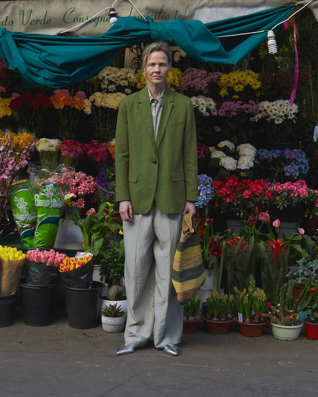 Adult posing in front of flower stand, wearing a green blazer, gray pants, and metallic shoes, holding a striped tote bag.