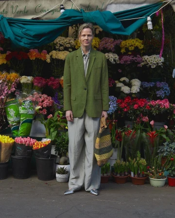 Adult posing in front of flower stand, wearing a green blazer, gray pants, and metallic shoes, holding a striped tote bag.