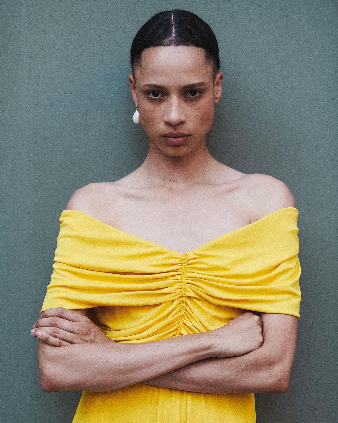 Elegant woman in yellow off-the-shoulder dress poses against a muted green wall, showcasing fashion and style with a serious expression.