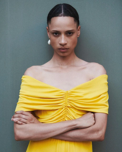 Elegant woman in yellow off-the-shoulder dress poses against a muted green wall, showcasing fashion and style with a serious expression.