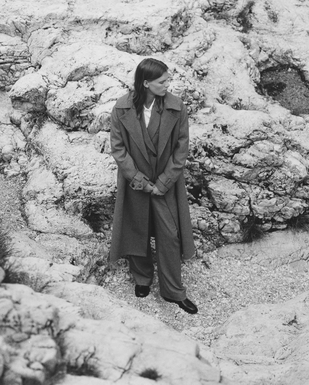 An adult woman in a coat and pants stands on rocky terrain. The greyscale image shows the woman looking to the side with a neutral expression.