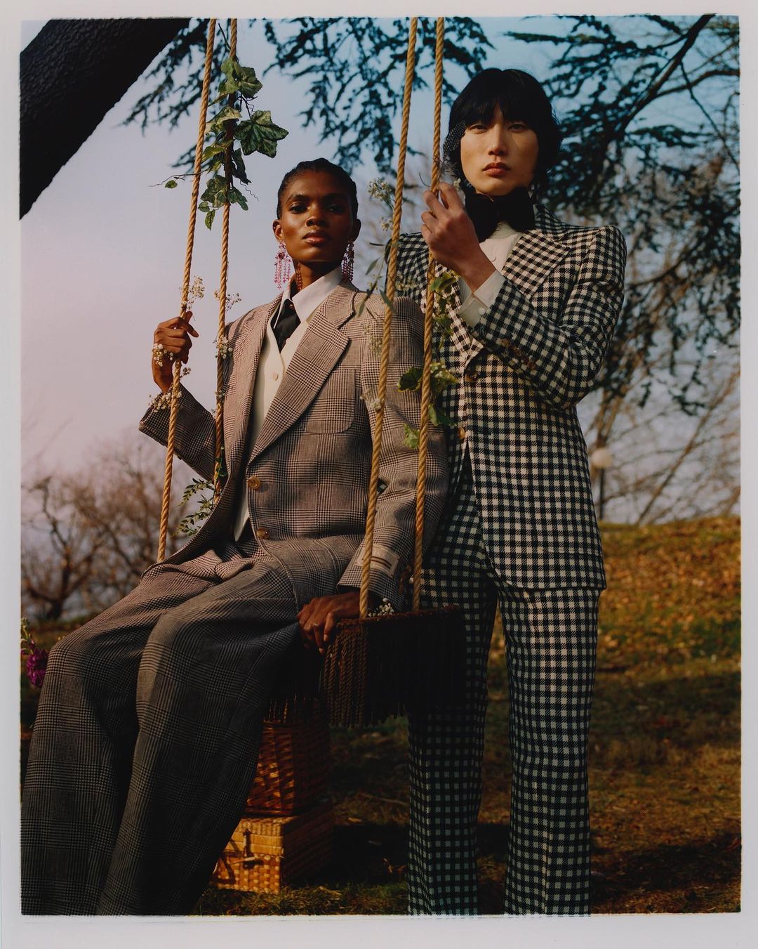 Two fashion models in a grassy outdoor setting. A Black woman is sitting on a swing and an Asian man is standing beside her.