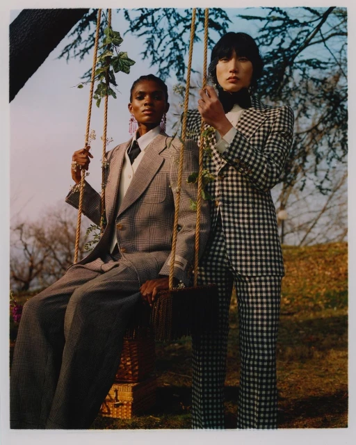Two fashion models in a grassy outdoor setting. A Black woman is sitting on a swing and an Asian man is standing beside her.