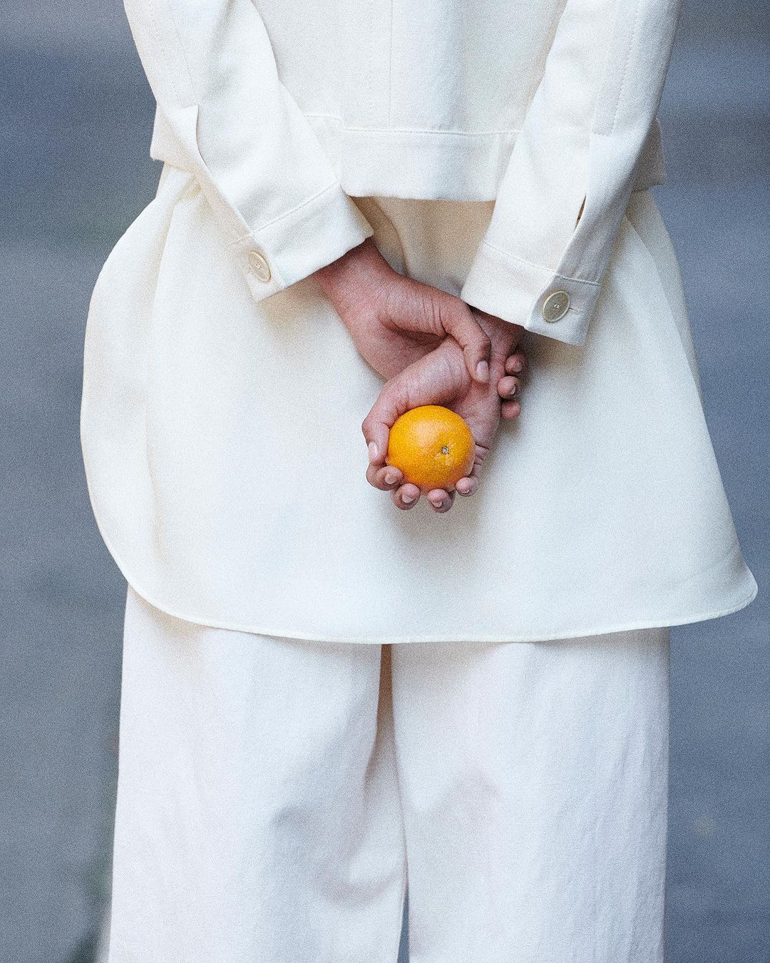 A woman in white jacket and pants holds an orange behind her back in a medium shot. Neutral grey studio backdrop. Fashionable and clean aesthetic.