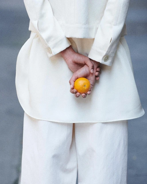 A woman in white jacket and pants holds an orange behind her back in a medium shot. Neutral grey studio backdrop. Fashionable and clean aesthetic.