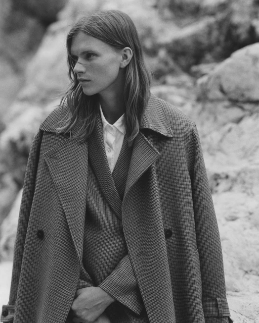 Monochrome portrait of a young adult with long hair, wearing a tailored suit and coat, posing outdoors against a rocky background.