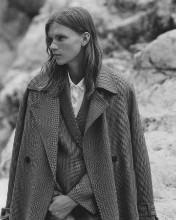Monochrome portrait of a young adult with long hair, wearing a tailored suit and coat, posing outdoors against a rocky background.