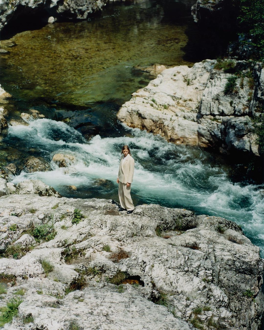 Woman in cream suit stands on rocky edge of river with rapid currents. Natural daylight, serene setting, portrait orientation.