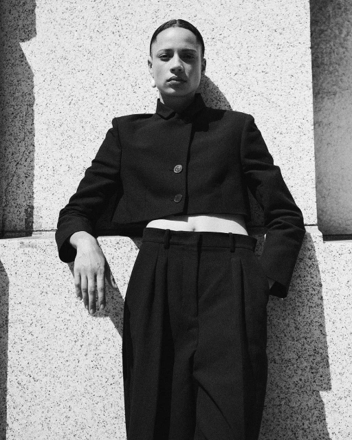 An androgynous person posing in a black cropped jacket and pants leans against a granite wall, bathed in hard sunlight.