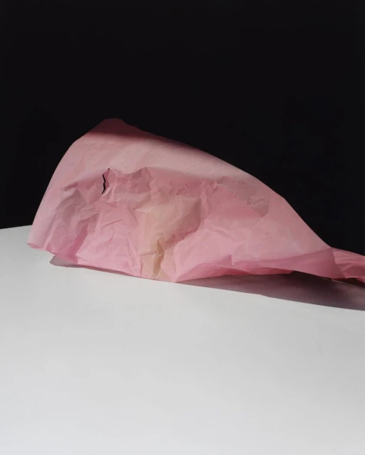 Minimalist still life photo of crumpled pink tissue paper on a white surface against a dark background, emphasizing textures and light.