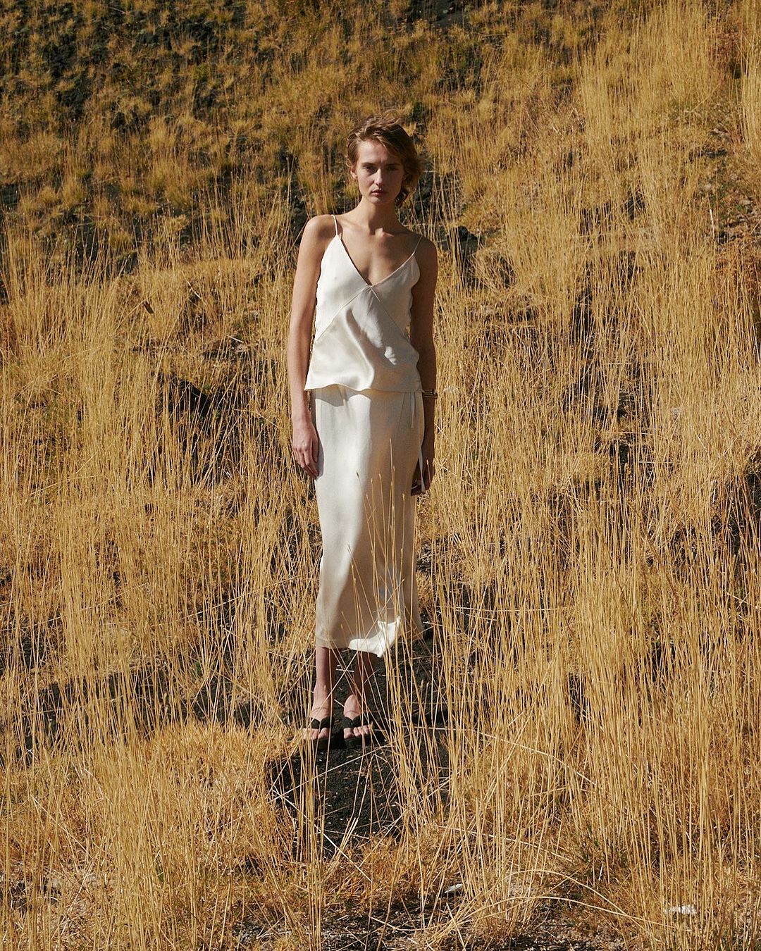 A fair-skinned woman stands in a cream-colored dress surrounded by tall, dry grass on a sunny day in an outdoor setting.
