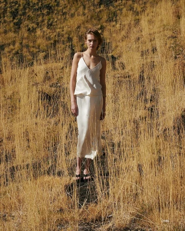 A fair-skinned woman stands in a cream-colored dress surrounded by tall, dry grass on a sunny day in an outdoor setting.